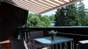 Un balcon doté d’un toit en lattes de bois, d’une table en verre, de chaises en osier et d’une plante en pot.