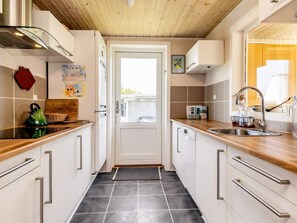 A narrow kitchen with a wooden ceiling, white cabinets, a stainless steel sink, and a door leading outside. - 4 Star Holiday Home in Hadsund (Hadsund)