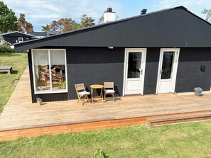 A modern black building with a wooden deck, two doors, and a large window overlooking a grassy area.