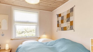 A bedroom with a bed, a window with blinds, a wall-mounted quilt, a bedside table with a lamp, and a wall-mounted light fixture. - 4 Star Holiday Home in Hadsund (Hadsund)