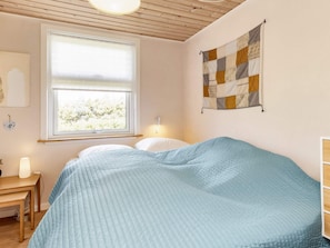 A bedroom with a bed, a window with blinds, a wall-mounted quilt, a bedside table with a lamp, and a wall-mounted light fixture. - 4 Star Holiday Home in Hadsund (Hadsund)