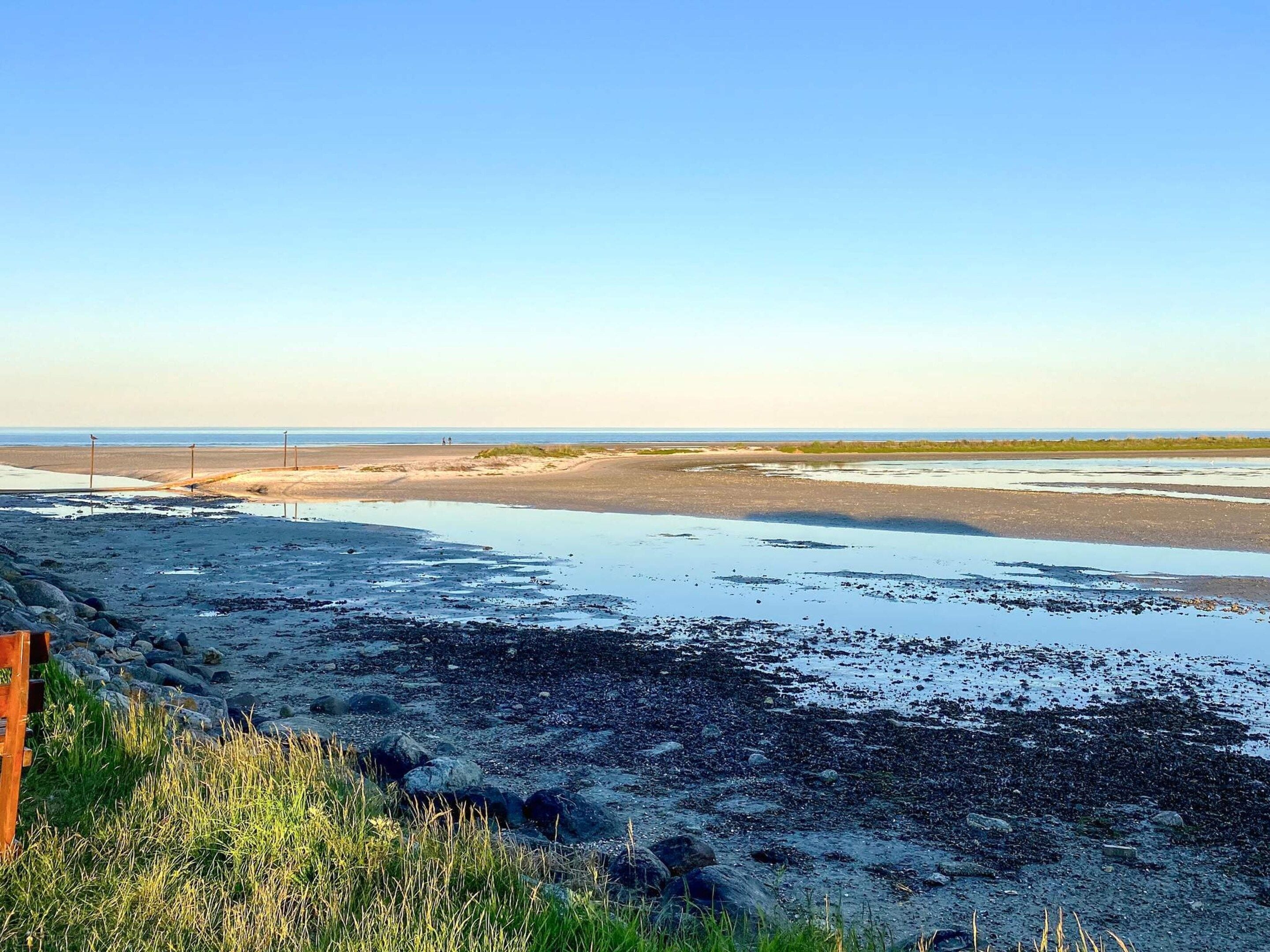 A coastal landscape with a sandy beach, rocky foreground, and a clear sky.