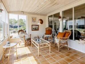 A sunlit room with wicker furniture, a tiled floor, and large windows offering a view of the outdoors.