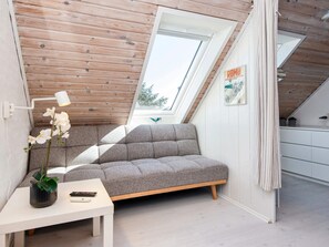 Dormitorio tipo loft con techo inclinado, un sofá gris, una mesita con lámpara y una ventana con vistas a árboles.