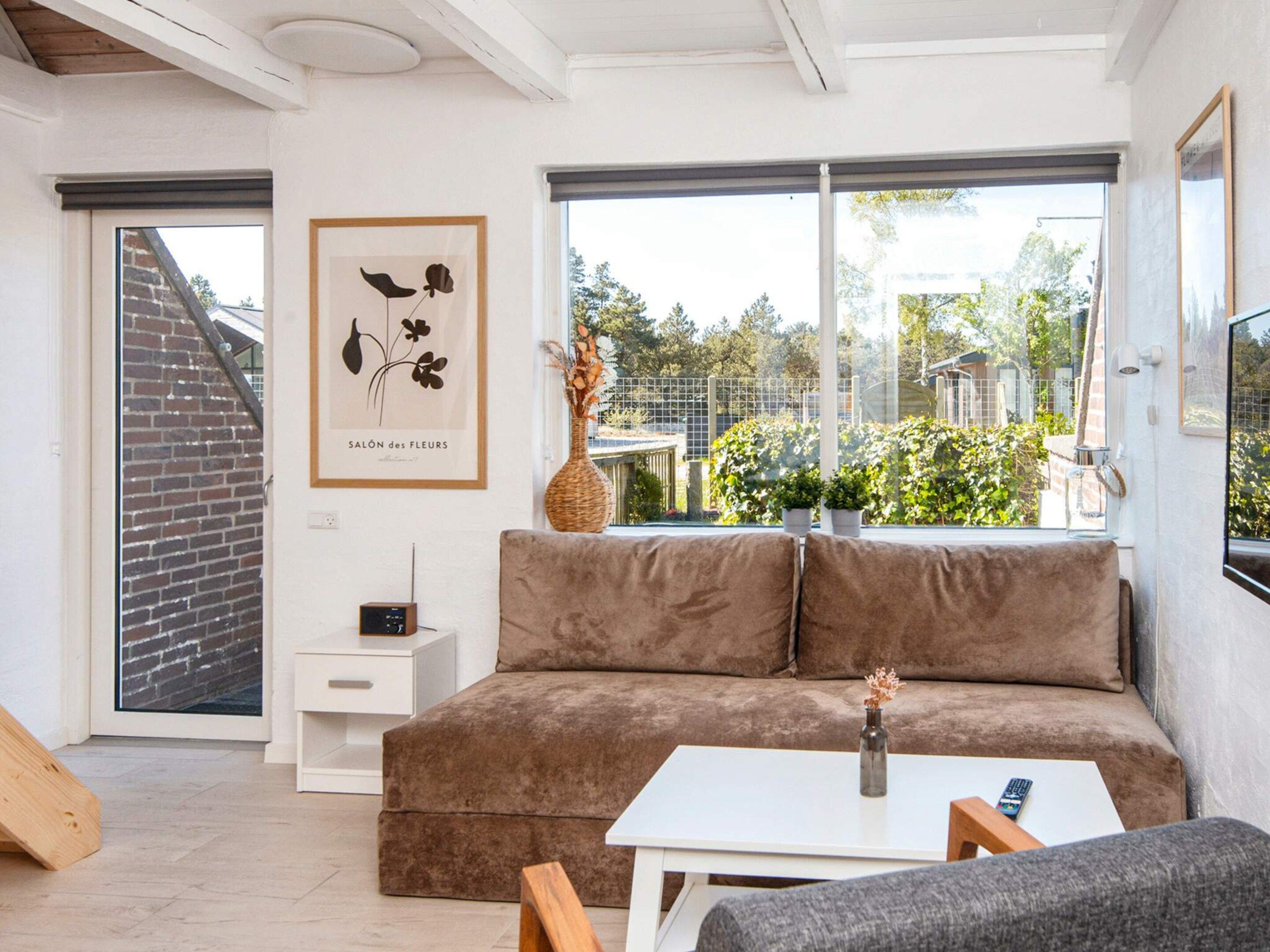 Ein modernes Wohnzimmer mit einem braunen Sofa, einem weißen Beistelltisch, einem gerahmten Kunstwerk und Blick ins Grüne.