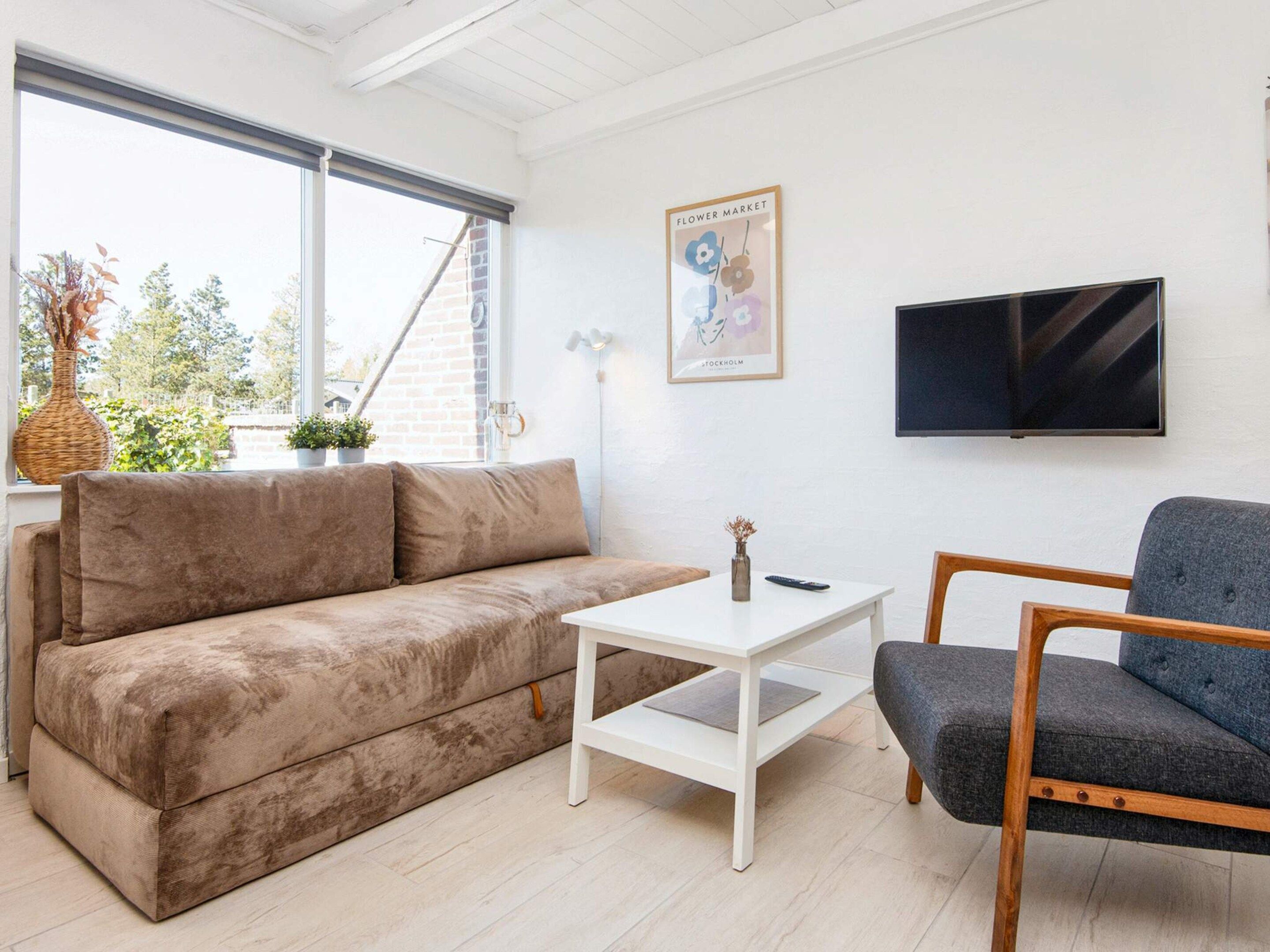A living room with a sofa, a coffee table, and a chair. There is a TV mounted on the wall and a window with a view of the outside.