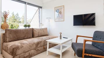 A living room with a sofa, a coffee table, and a chair. There is a TV mounted on the wall and a window with a view of the outside.