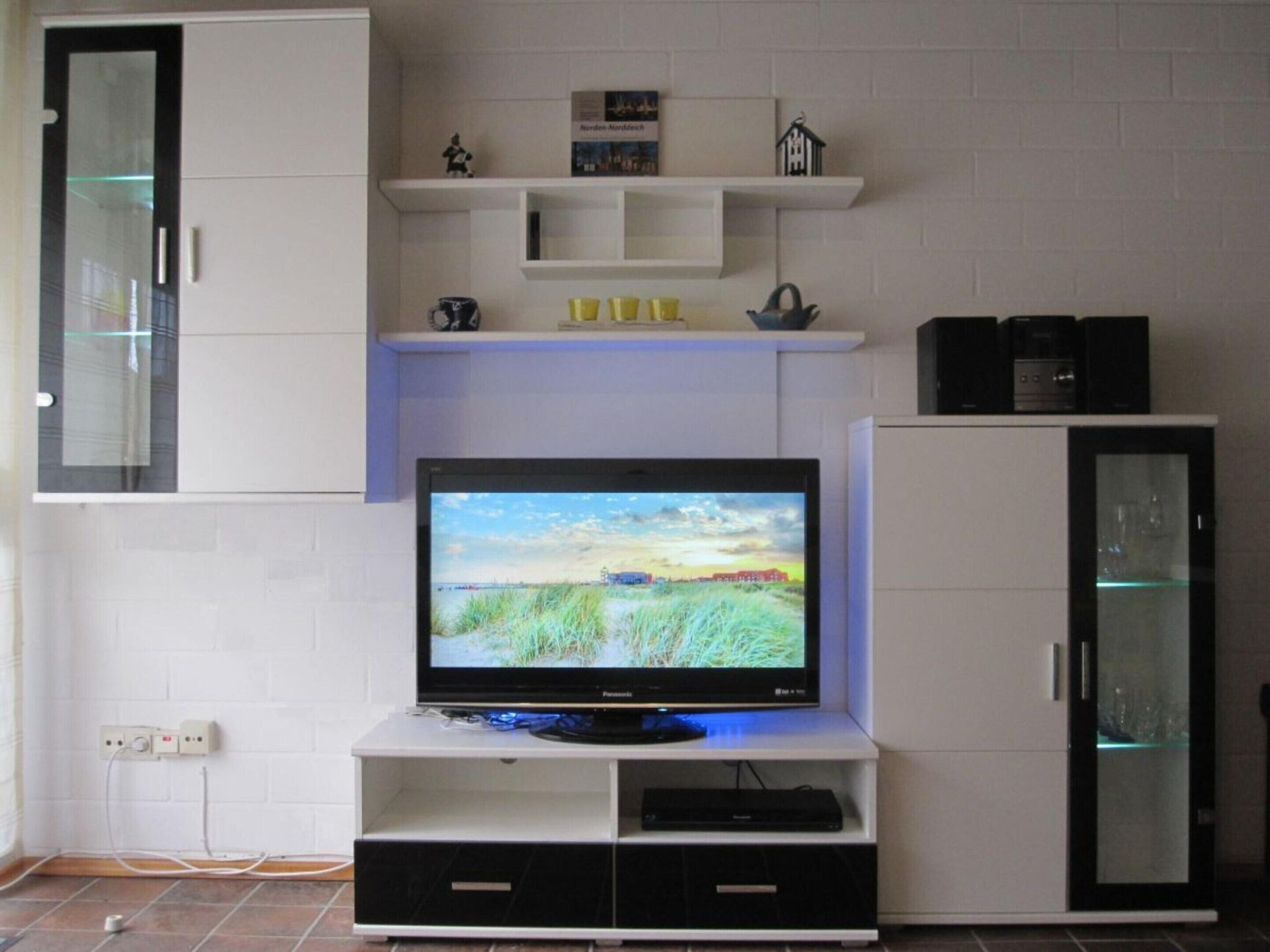Un salon moderne avec un téléviseur à écran plat posé sur un meuble blanc, un buffet avec des portes en verre et une étagère ornée d’objets décoratifs.