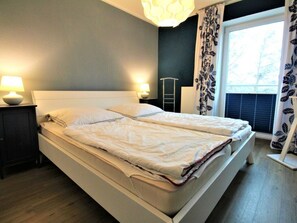 A neatly made bed with white bedding in a room with a dark wall, a window with blinds, and two bedside lamps. - Residence "hohen-lith" 2-14 (Cuxhaven)