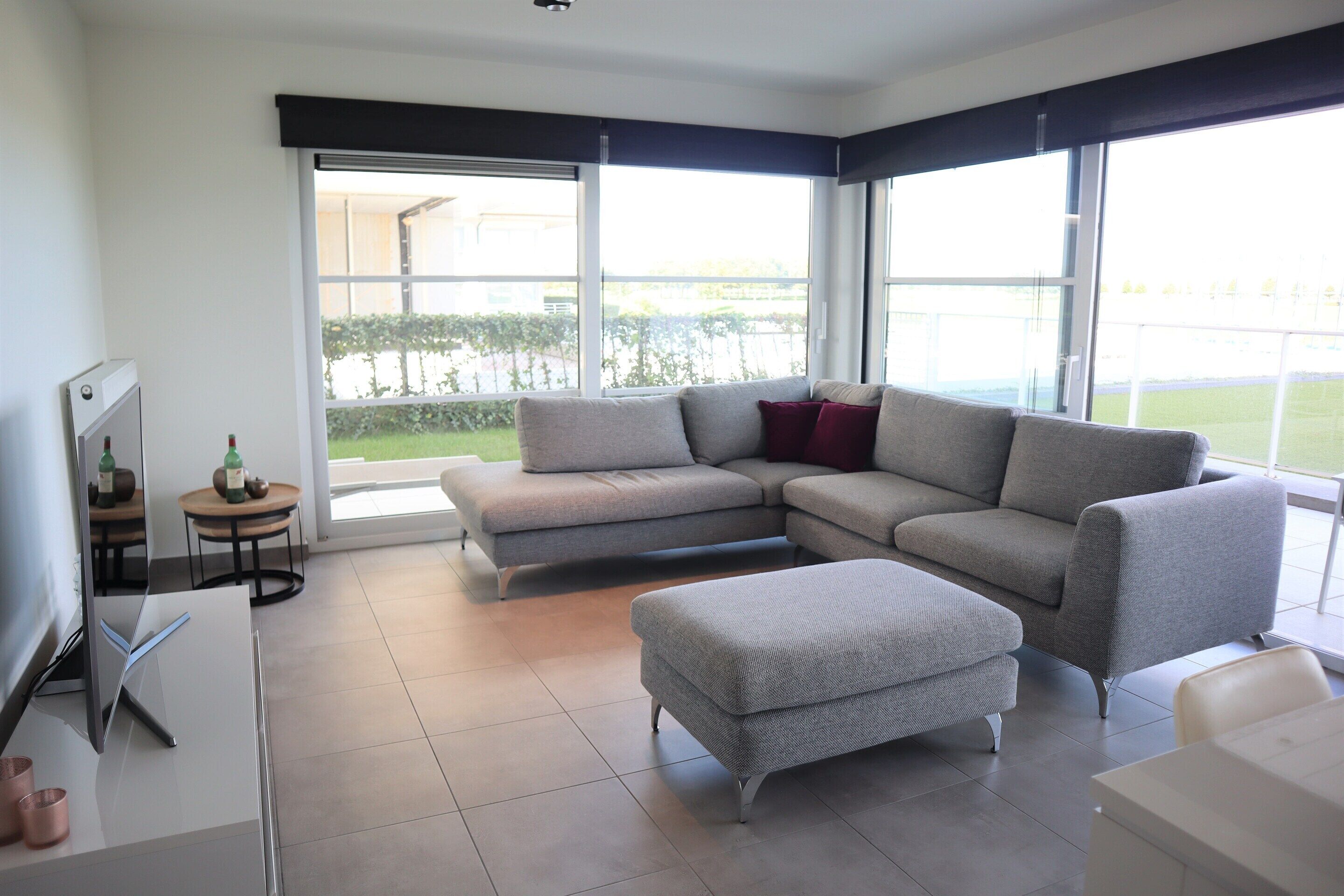 A modern living room with a grey sectional sofa, a matching ottoman, and a small table with a bottle and glasses. Large windows provide a view of the outdoors.