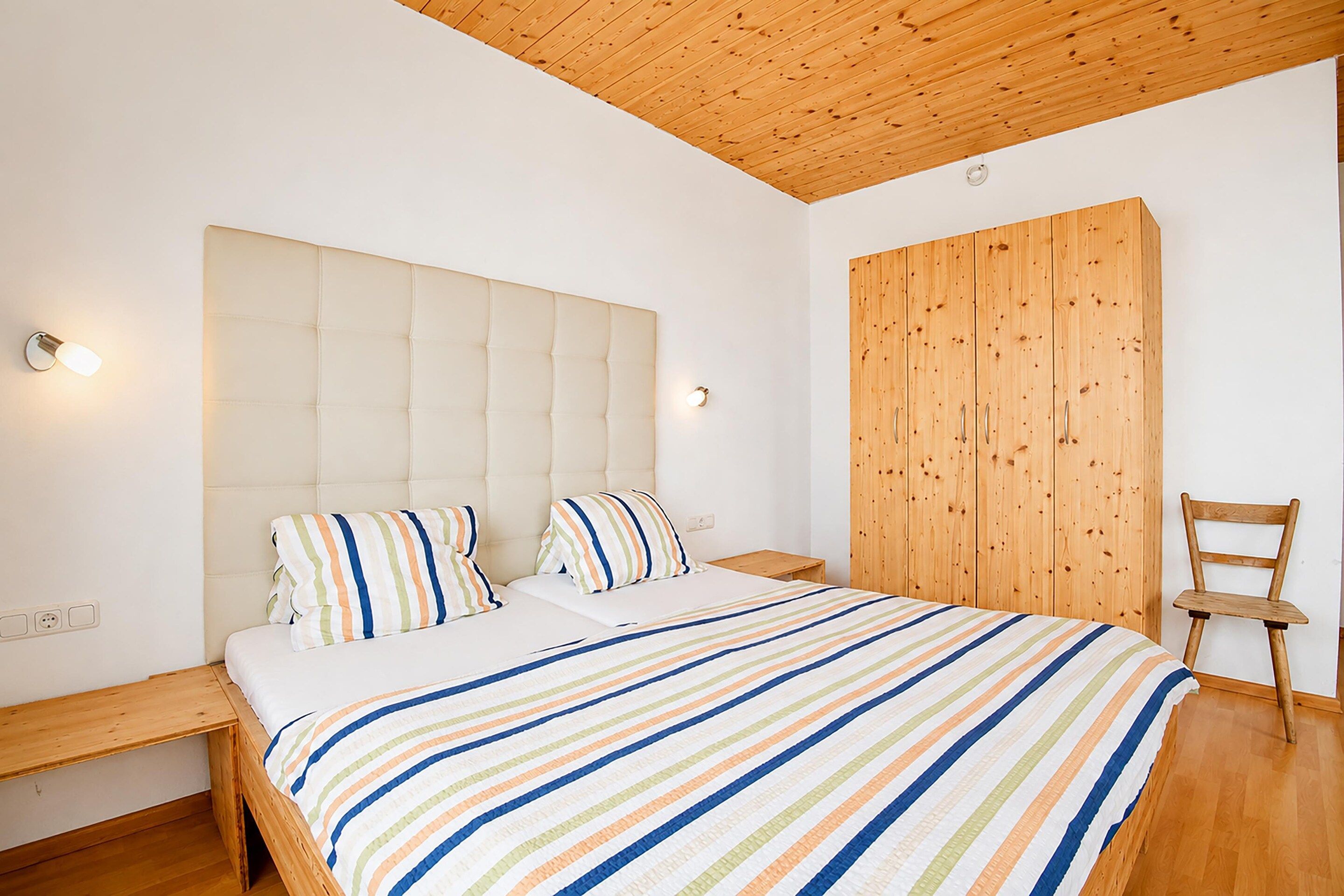 A bedroom with a large bed, a wooden wardrobe, a wooden chair, and a nightstand with a lamp.