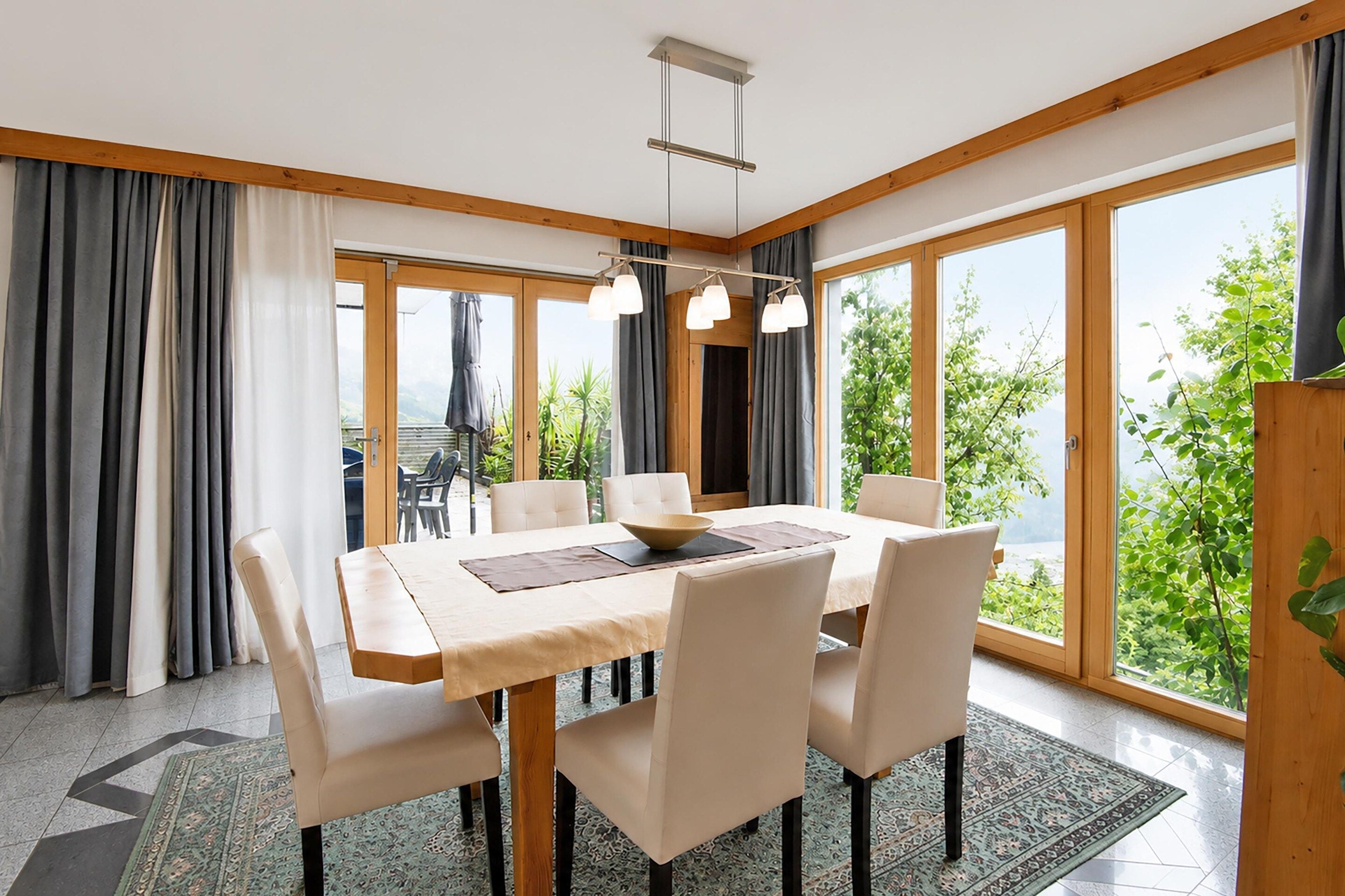 A dining area with a table set for six, large windows, and a view of greenery.
