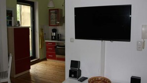 A living room with a flat-screen TV mounted on the wall, a small table with a bowl, a chair, and a kitchen area with a refrigerator, stove, and cabinets. - Holiday Apartment Casabrema (Bremen)