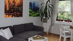 A living room with a grey sofa, a small white table, and a potted plant. There are two wall-mounted paintings and a window with curtains. - Holiday Apartment Casabrema (Bremen)