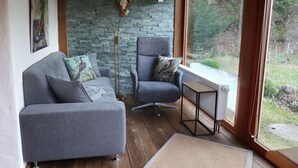A cozy living room with a grey sofa and armchair, a stone wall, and a large window offering a view of the outdoors. - Waterval in het Huis Boshoek (Aschau im Chiemgau)
