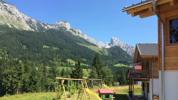 A wooden cabin with a small play area, including a swing set and a slide, set against a backdrop of green hills and mountains. - Huttenzauber 2 Comfortable Holiday Residence (Annaberg-Lungoetz)