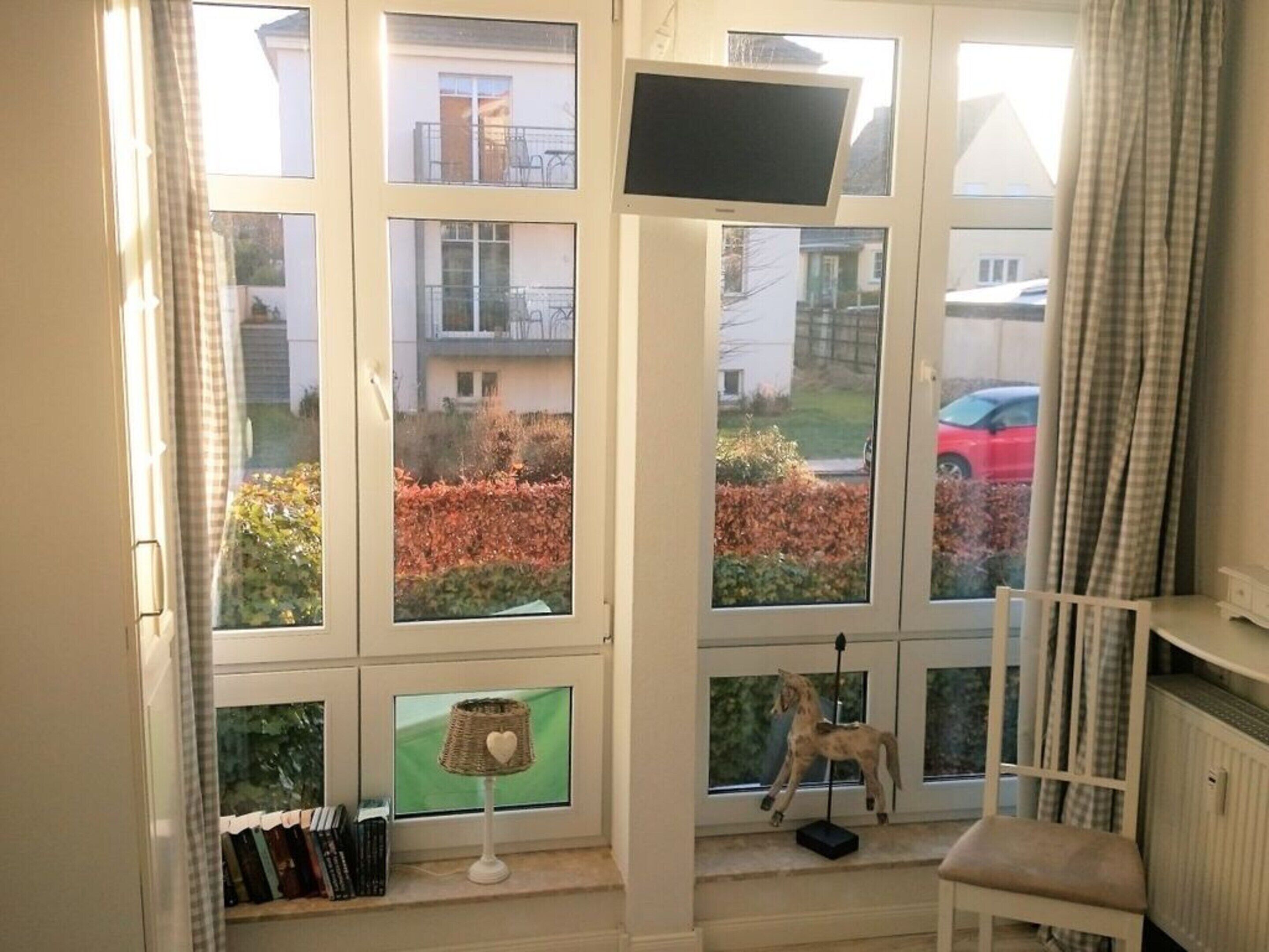A room with a large window, a television, a small table with a lamp, and a chair.