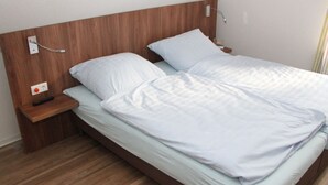 A single bed with a wooden headboard, a bedside table with a remote control, and a power outlet.