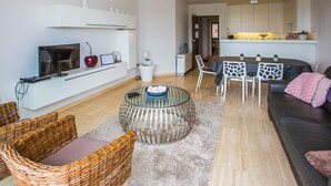 Una sala de estar moderna con televisor, computadora portátil y sillas de mimbre. Hay una mesa de centro de vidrio y un sofá con una manta rosa.