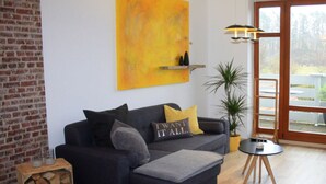 A modern living room with a dark grey sofa, a yellow abstract painting, a wooden side table, and a potted plant. - Living Lust 3 in Malchow (Malchow)