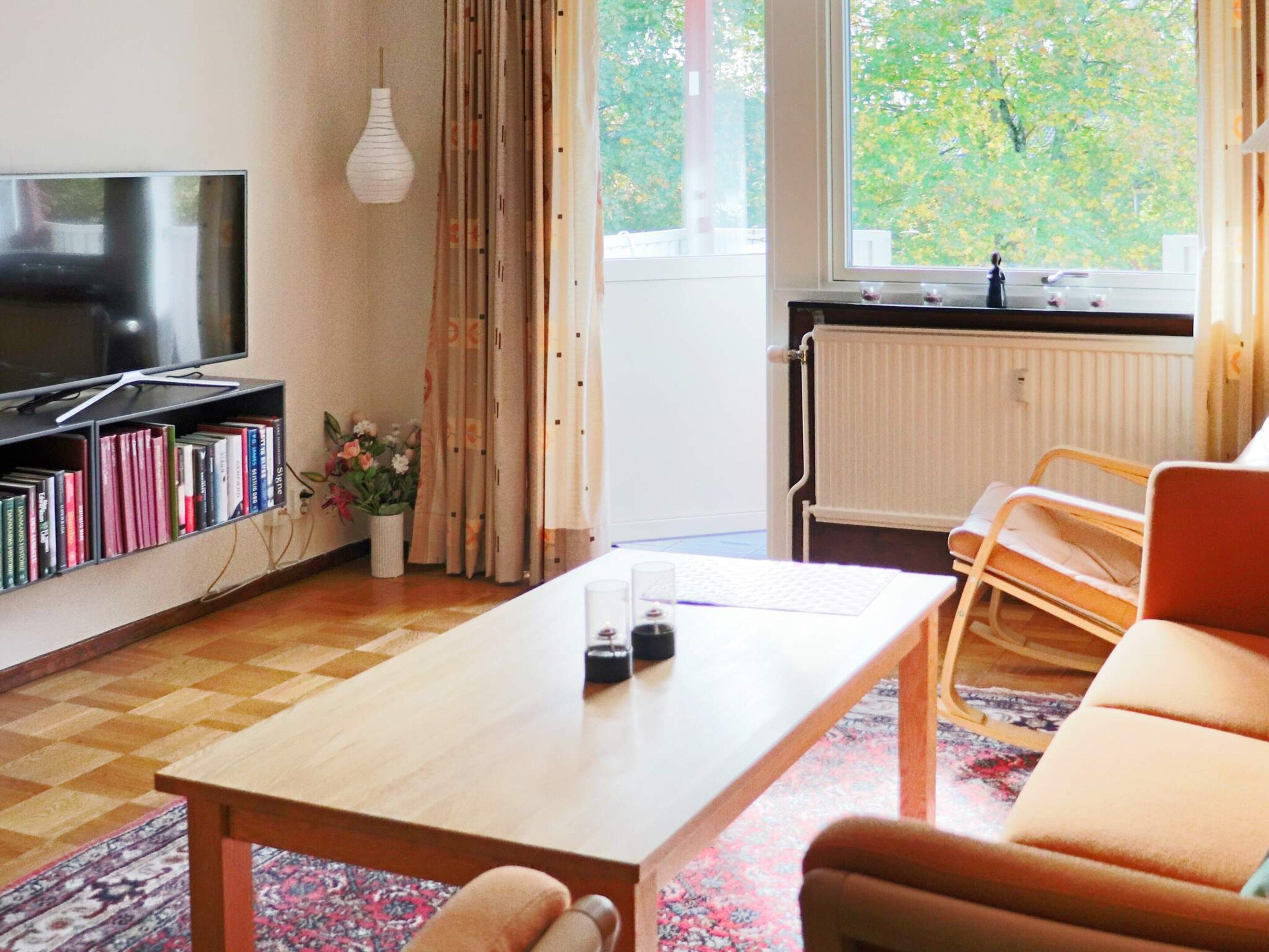 Olohuone, jossa on televisio, kirjahylly, sohvapöytä ja ikkuna, josta avautuu näkymä puita kohti.