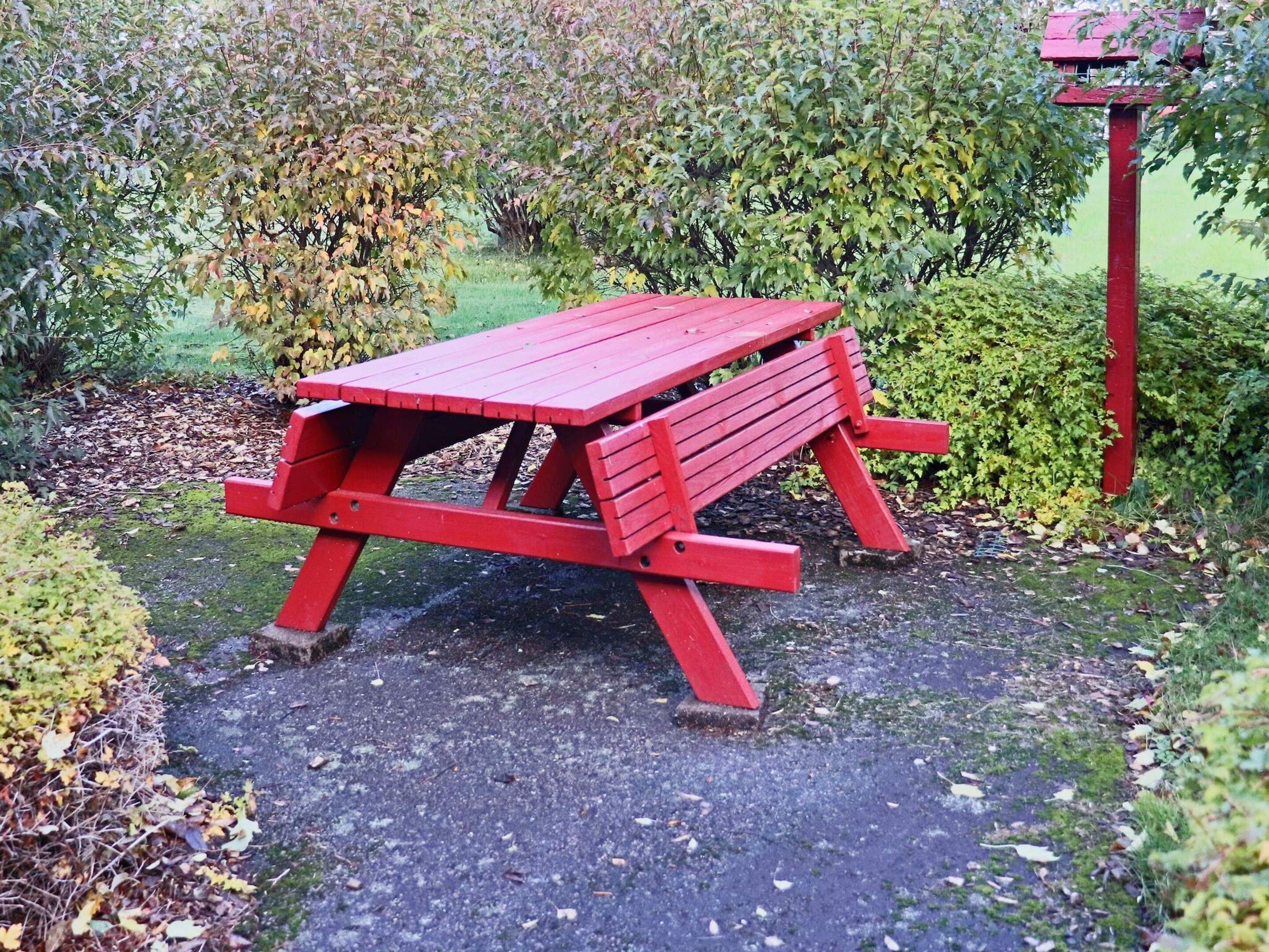 A red picnic table in a garden with a red post and a birdhouse in the background.