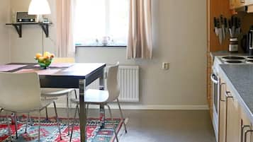 A kitchen with a dining table, chairs, a bowl of fruit, a hanging light, and a window with curtains.