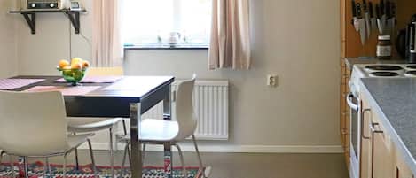 A kitchen with a dining table, chairs, a bowl of fruit, a hanging light, and a window with curtains.