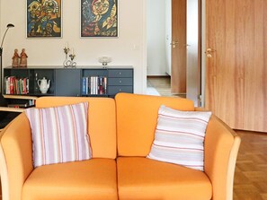 A living room with an orange sofa, two pillows, a bookshelf, and two abstract paintings on the wall. - 4 Person Holiday Home in Hyltebruk (Hyltebruk)