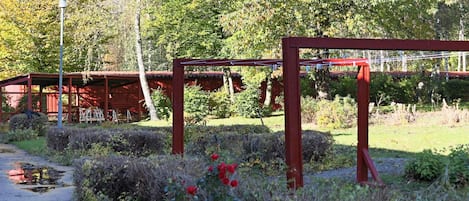 Un sendero de jardín con una estructura metálica roja, rodeado de vegetación y un edificio rojo al fondo.