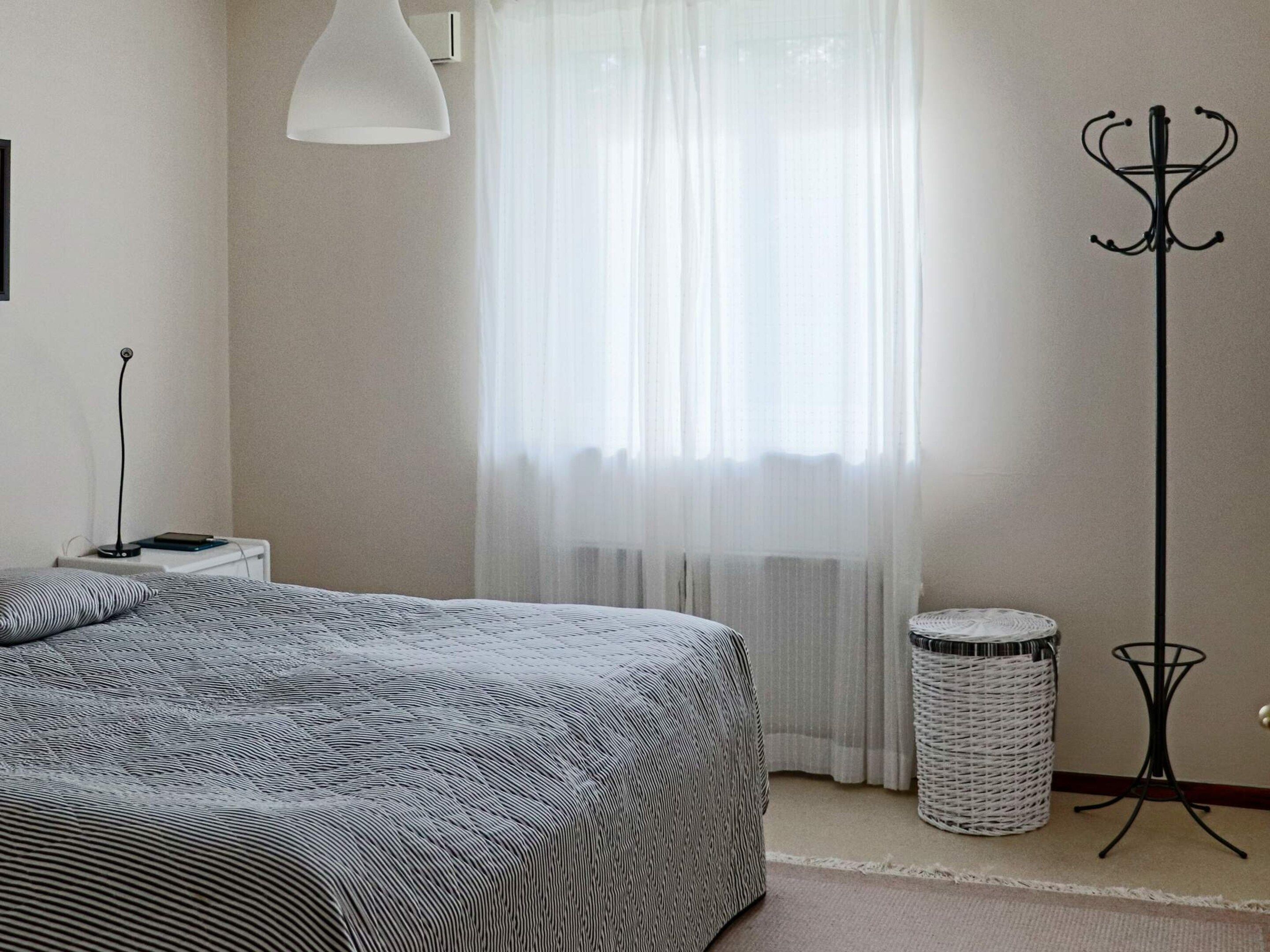 A bedroom with a bed, a bedside lamp, a floor lamp, a waste basket, and a window with sheer curtains.