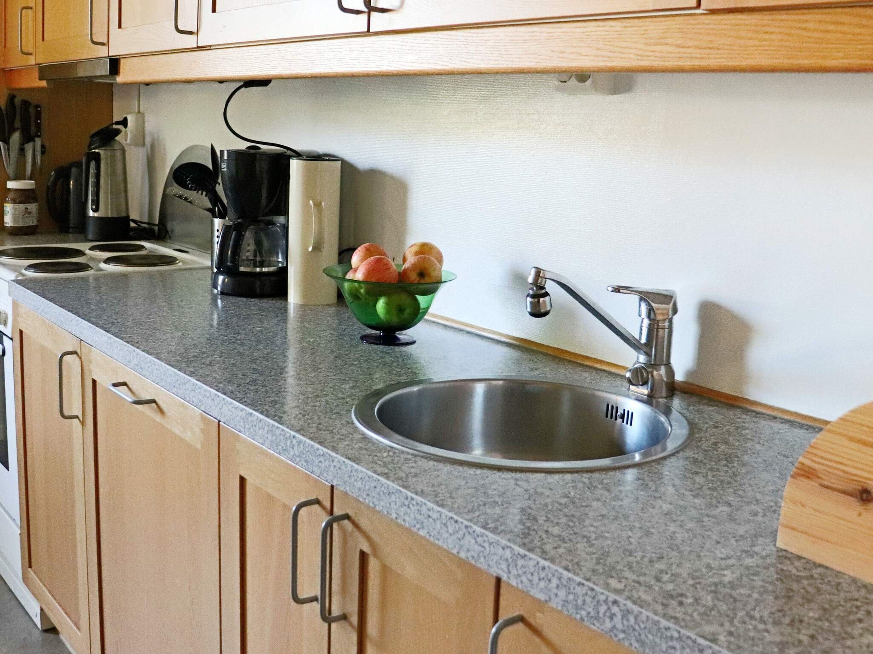 A kitchen with a granite countertop, a stainless steel sink, wooden cabinets, a coffee maker, and a bowl of fruit.