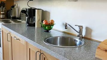 A kitchen with a granite countertop, a stainless steel sink, wooden cabinets, a coffee maker, and a bowl of fruit.