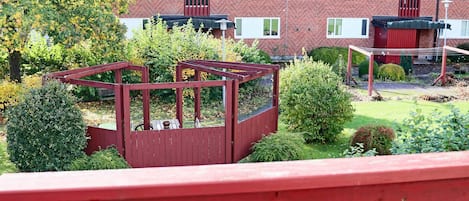 Un barrio residencial con una terraza de madera roja, una casa de ladrillo con techo de tejas y un jardín con arbustos y árboles variados.