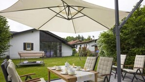 An outdoor patio with a table set for tea, surrounded by cushioned chairs, under a large umbrella. - Kaiserblick Comfortable Holiday Residence (Oberaudorf)