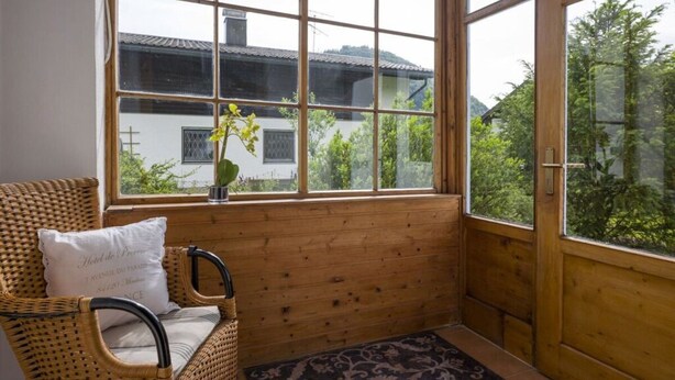 A cozy room with a wicker chair, a small table with a vase, and a view of a white building through large windows. - Kaiserblick Comfortable Holiday Residence (Oberaudorf)