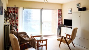 A modern living room with a sofa, armchairs, a coffee table, and a TV. There is a sliding glass door leading to a balcony. - Apartment in Nieuwpoort With sea View (Nieuwpoort)