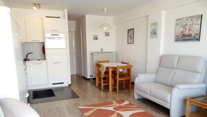 A modern living room with a sofa, a dining table with chairs, a kitchen area with a refrigerator, and a patterned rug. - Apartment in Nieuwpoort With sea View (Nieuwpoort)