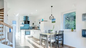 A modern kitchen with white cabinets, a dining area with a wooden table and chairs, and a large window with a view of greenery. - Nitschke - Vakantiewoning Watt (Aurich)