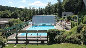 A swimming pool with a covered area, surrounded by greenery and a few buildings.