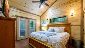 Une chambre dans une cabane en bois, avec un lit, un ventilateur de plafond, une fenêtre donnant sur les arbres et une porte menant à l’extérieur.