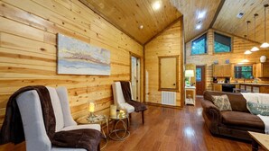 L’intérieur d’une cabane en rondins, avec un coin salon accueillant, des murs en bois et une cuisine visible à travers une porte ouverte.