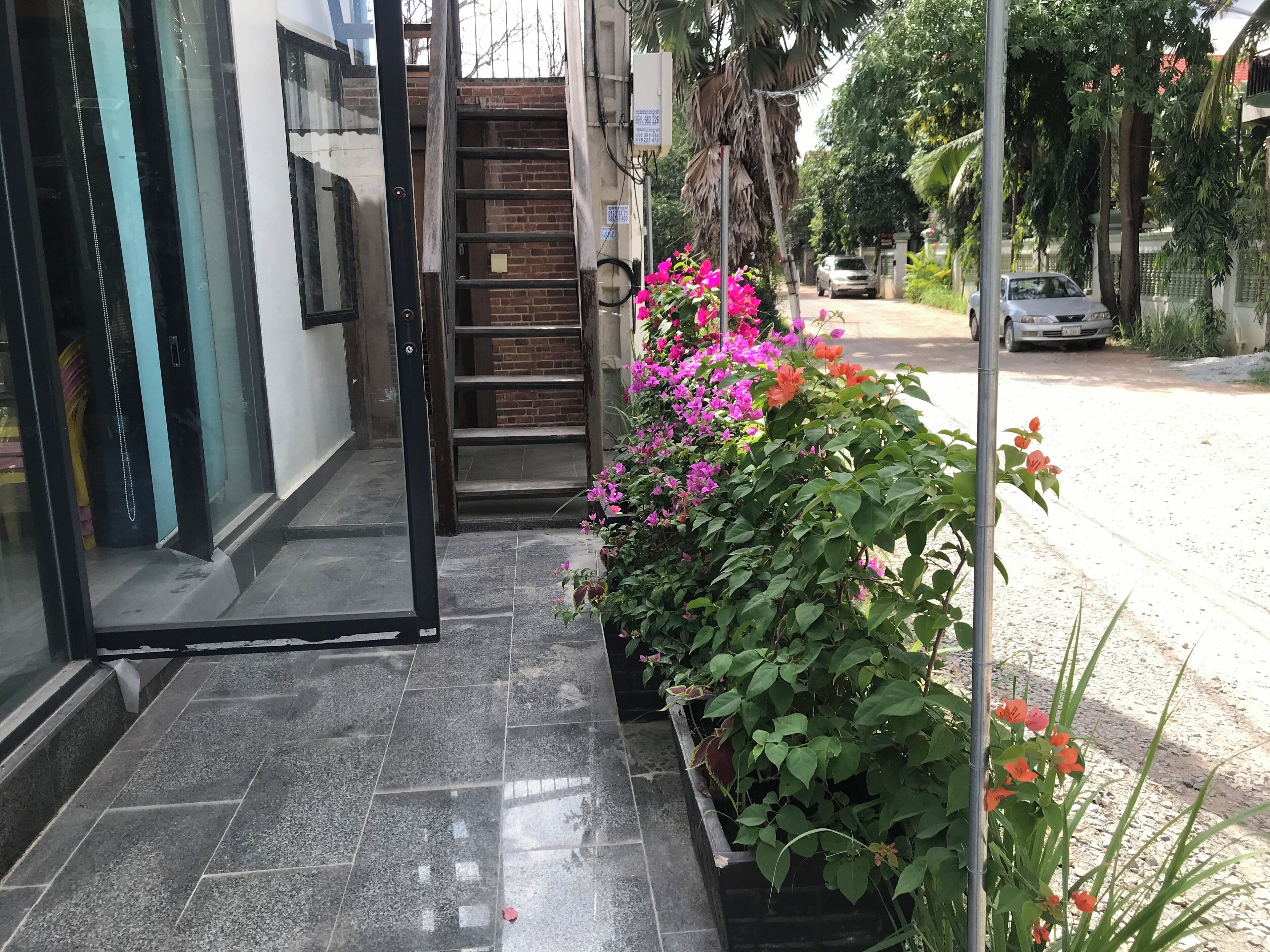 A hotel entrance with a glass door, a tiled floor, and a small garden with pink flowers.