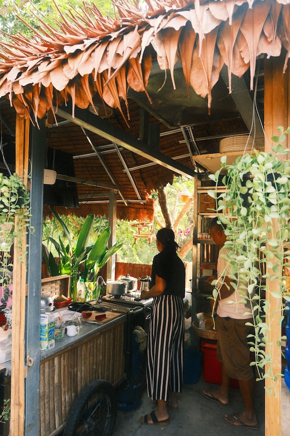 A rustic outdoor kitchen with a wooden counter, hanging plants, and a thatched roof. - Madam Sokha Homesteading (Siem Reap)