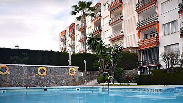 A swimming pool with a stone wall and lifebuoys, surrounded by a multi-story building with balconies.