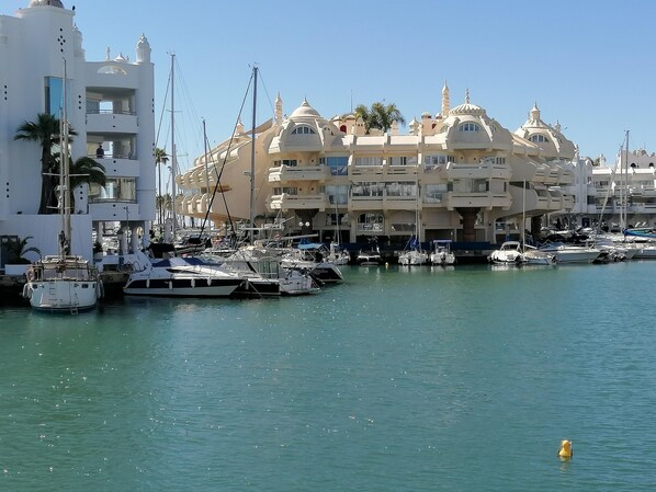 A marina with multiple boats docked, a white building with balconies, and a clear blue sky.