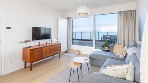 Un salon moderne doté d’un grand téléviseur, d’un meuble en bois, d’un canapé gris et offrant une vue sur la mer grâce à de grandes fenêtres.