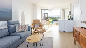 Un salon moderne avec un canapé, un fauteuil et une table basse. On aperçoit à l’arrière-plan un coin repas avec des chaises et une table.