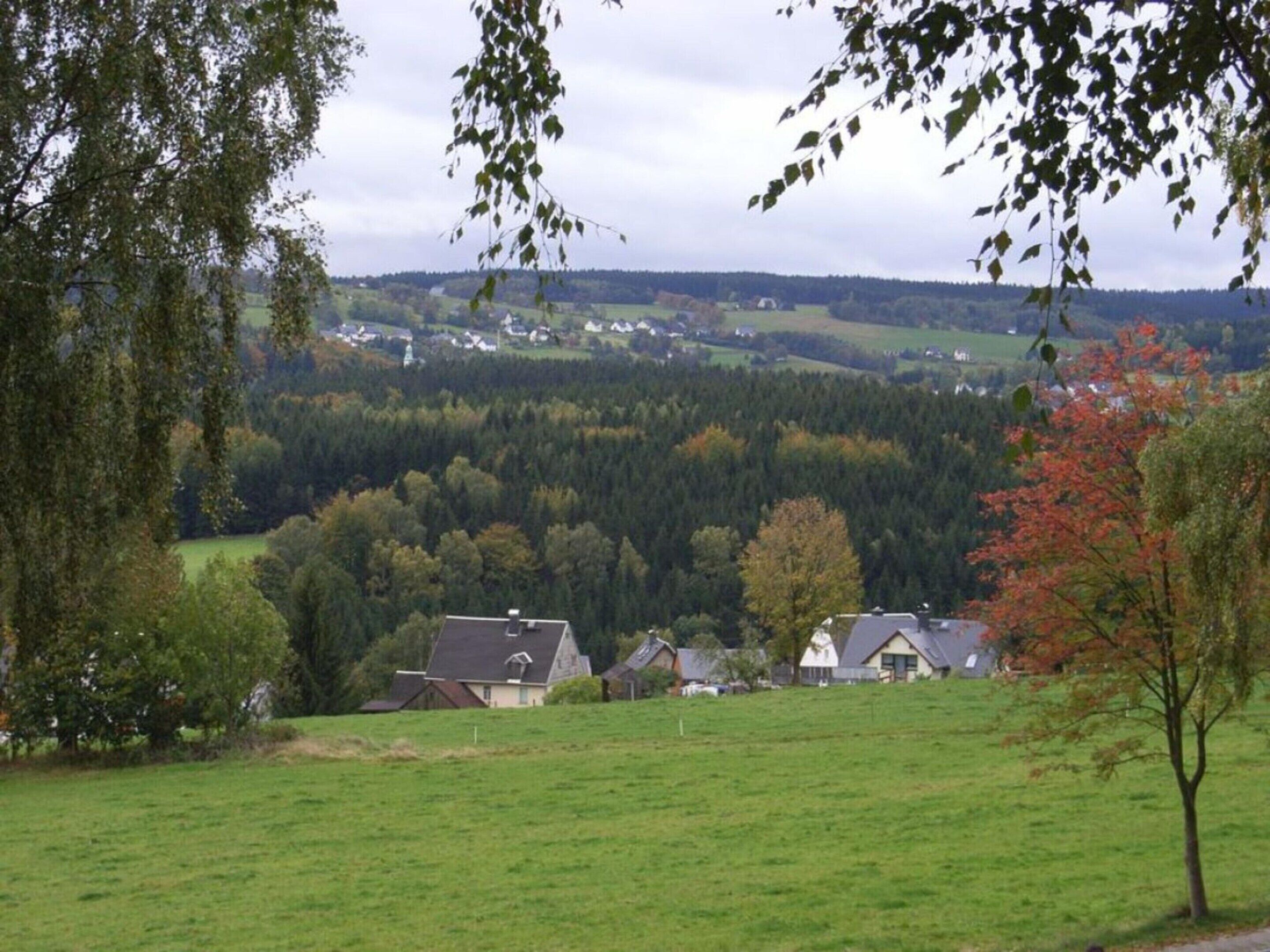 Ländliche Landschaft mit Häusern auf einem Hügel, umgeben von grünen Feldern und Bäumen.