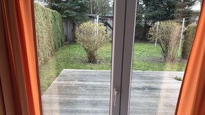 A view through a sliding glass door into a backyard with a wooden deck, a small bush, and a hedge. - Fabian Comfortable Holiday Residence (Wieck auf dem Darss)